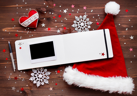 Christmas and New Year background with felt heart, red Santa's hat, notepad with pen, empty photo frame and christmas decorations - stars, silver sparkling snowflakes and confetti on wooden table. Place for your text.の写真素材