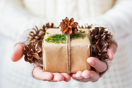 Woman in knitted sweater holding a present. Gift is packed in craft paper with pine cones and tied with rough rope. Hands full of pine cones. Example of DIY way to wrap a present.の写真素材
