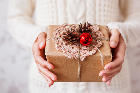 Woman in knitted sweater holding a present. Gift is packed in craft paper with pine cones, crismas decorative red ball. Hand made crocheted tiny napkin on box, tied with rough rope. Example of DIY way to wrap a present.の写真素材
