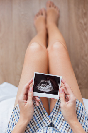 Woman in shirt sitting on bed with Ultrasound photo of baby in hands. Woman is expecting baby. Cozy happy morning at home.の写真素材