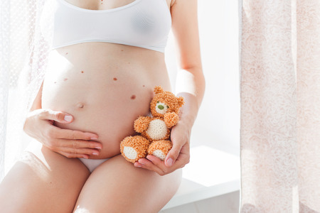 Pregnant woman in white underwear with knitted toy teddy bear. Young woman expecting a baby. Cozy happy background in sunny morning.の写真素材