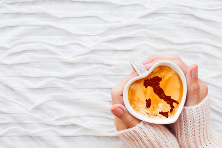 Woman holds a cup of hot coffee with cinnamon silhouette of Italy. Winter wanderlust and Valentine's Day fabric background.の写真素材