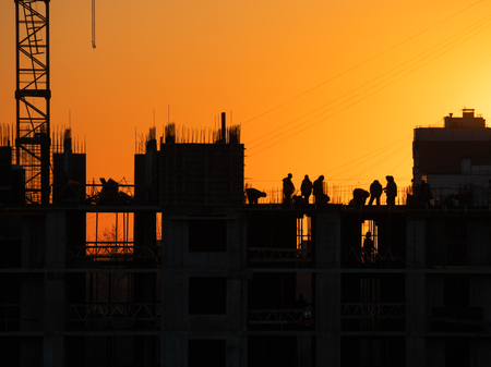 Construction of residential building. Builders go on the unfinished floor with protruding fittings. Silhouettes of workers on the background of an orange sunset.の写真素材