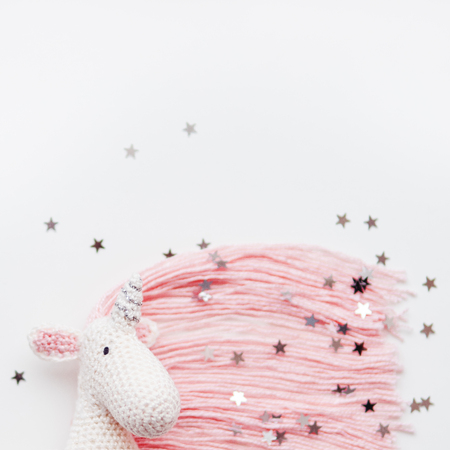 Cute fairy unicorn with a pink mane and a tail made of threads. Crocheted hand made toy on white background with silver stars confetti. Trendy creature, symbol of magic and miracles. Place for text.の写真素材