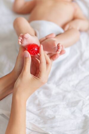 Mother holds newborn baby's feet. Tiny fingers and red massage ball in woman's hand. Cozy morning at home.の写真素材