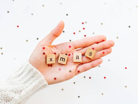Word XMAS on palm. Woman in knitted sweater holding wooden letters with stars confetti. Christmas and New Year background.の写真素材