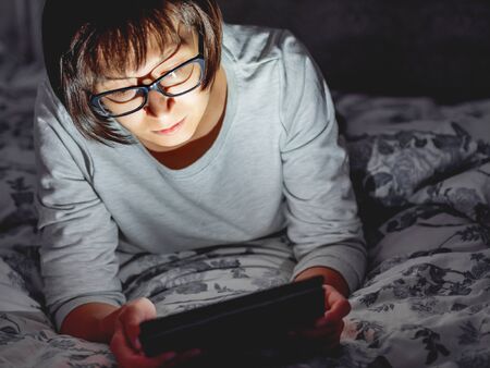 Woman in glasses are lying in bed with tablet in dark room. She is touching screen. Night bedtime.の写真素材