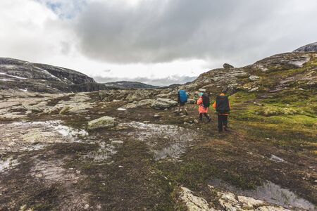 Tourists with backpacks walk across a rocky plain to the famous landmark - Troll tongue rock or Trolltunga. Cold and windy summer in Norway.の写真素材