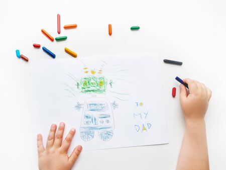 Toddler draws greeting card with funny robot for Father's day or Daddy's Birthday. Kid uses wax crayons. Top view on child's hands and pencils on white background.の写真素材