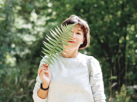 Young woman is hiding her eyes with fern leaf. Symbol of life, tranquility and unity with nature. Summer in forest.の写真素材