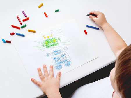 Toddler draws funny robot. Kid uses wax crayons. Top view on child's hands and pencils on white background.の写真素材
