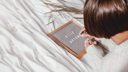 Woman in white knitted sweater typing HELLO AUTUMN on letter board. Crumpled textile background with dried grass. Season greeting banner with copy space. Sunny, warm and cozy home.の写真素材