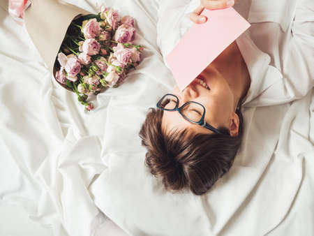 Young smiling woman is lying on crumpled bed sheet. She received bouquet of lilac colored roses and letter in pink envelope. Cute present for Valentine Day or Happy Birthday surprise.の写真素材