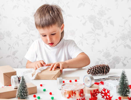 Kid wraps handmade Christmas presents in craft paper with colorful pompons and snowflake ribbons. Child prepares gifts for New Year celebration. Peaceful leisure activity before winter holidays.の写真素材