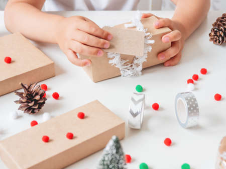 Kid wraps handmade Christmas presents in craft paper with colorful pompons and snowflake ribbons. Child prepares gifts for New Year celebration. Peaceful leisure activity before winter holidays.の写真素材