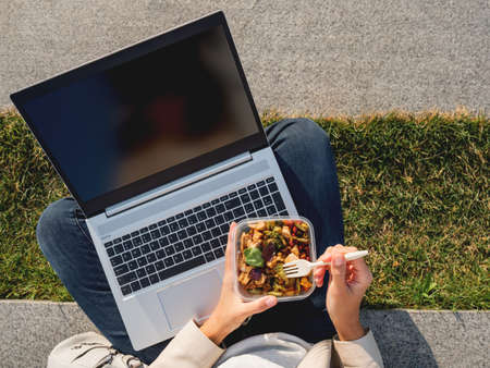 Woman sits on park bench with laptop and take away lunch box. Healthy bowl with vegetables. Casual clothes, urban lifestyle of millennials. Healthy nutrition.の写真素材