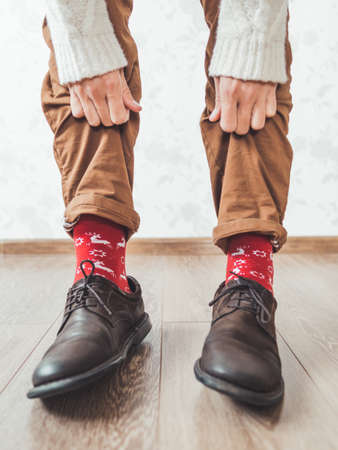 Young man pulls up leg of his chinos trousers to show bright red socks with reindeers on them. Scandinavian pattern. Winter holiday spirit. Casual outfit for New Year and Christmas celebration.の写真素材