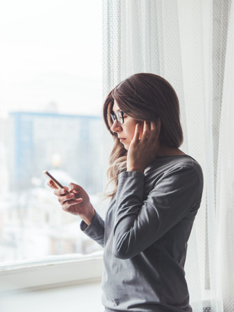 Thoughtful woman with eyeglasses looks at her smartphone. Information in online media. Wireless...