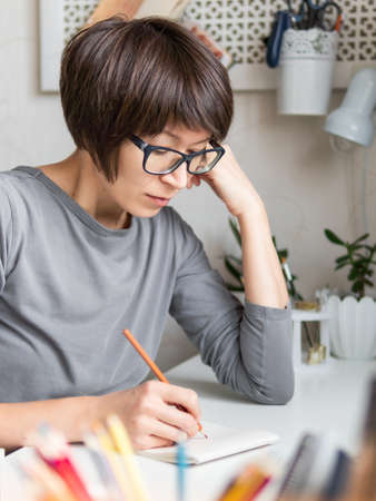 Woman with short hair cut is drawing in notebook. Calming hobby, antistress leisure. Artist at work. Cozy workplace.の写真素材