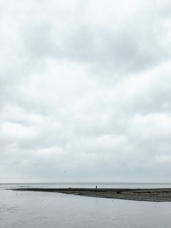 Alone man walking on pebble beach at windy weather. Gray cloudy sky upon seaside.