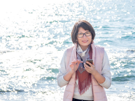 Smiling woman texting somebody on smartphone. Active recreation on seaside. Vacation on ocean coast. Turquoise water behind woman with happy smile.の写真素材
