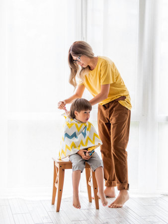 Mother prepares to cut her son's hair by herself. Little boy sits, covered with cloth, and holds...