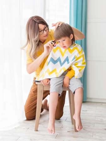 Mother cuts her son's hair by herself. Little boy sits, covered with cloth. New normal in case of quarantine and lockdown. Beauty and self care at home lifestyle.の写真素材