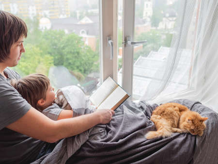 Father reads book to his son. Cozy family time on window sill with sleepy ginger cat. Dad, toddler boy and fluffy pet at home.の写真素材