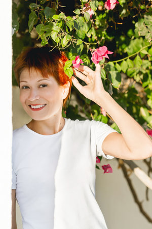 Smiling woman with short red hair poses with bougainvillea flowers. Summer vibes. Pretty female on hard sunlight. Vacation in country with tropical climate.の写真素材