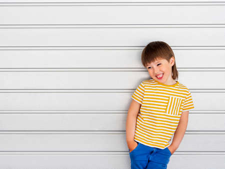 Portrait of smiling boy on white background with stripes. Cute kid stands hands in pockets near light gray wall. Child in yellow t-shirt and blue jeans laughs joyfully.の写真素材