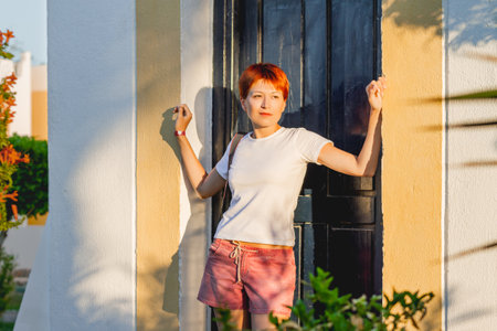 Young woman with short red hair stands near shabby door at sunset. Portrait of pretty female at hard sunlight. Vacation in tropical country.の写真素材