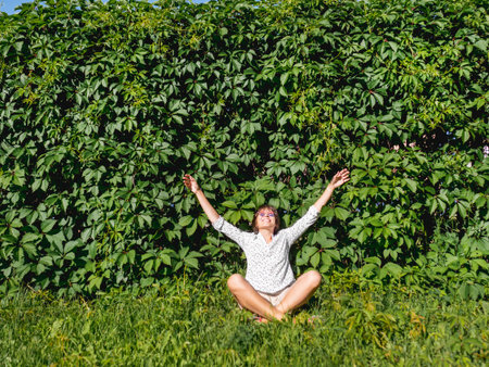 Wide smiling woman sits on grass lawn near wall all over covered with thick ivy. Natural background with green leaves and woman enjoying sunlight and warmth in urban park. Summer vibes.の写真素材