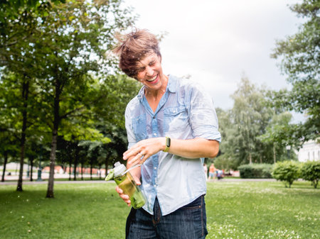 Laughing man douses himself with water from bottle. Staying cool in summer. Wet fun in urban park. Sincere emotions. Candid lifestyle.の写真素材