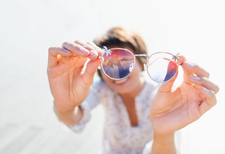 Woman with short hair looks through colorful sunglasses. Smiling woman at open wooden scene of urban...