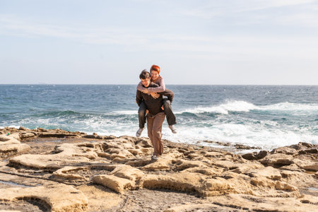 Happy couple on seaside. Man carries his girlfriend along rocky beach of Saint Julian area of Malta. Fun at sunny day.の写真素材