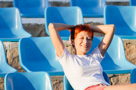 Young woman with short red hair sits relaxed in deserted open air audience. Summer vibes. Sport stadium with plastic blue seats.の写真素材