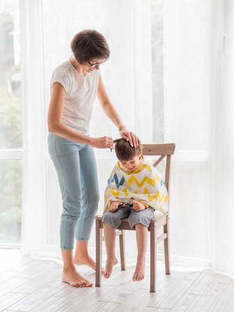 Mother cuts her son's hair by herself. Little boy sits, covered with cloth, and holds pair of scissorsの写真素材