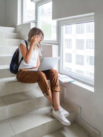 Thoughtful woman looks in notepad with handwriten notes. Student is working with laptop on stairs at university campus. Wireless technologies for education. Homework online. Using Internet to search for information.の写真素材
