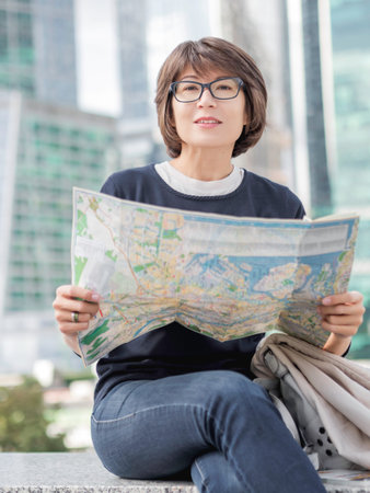 Smiling woman in eyeglasses is reading paper map sitting on background of buildings with glass...