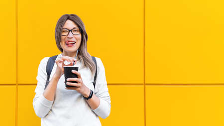 Wide smiling woman drinks coffee with black straw. Portrait of happy female with tasty cappuccino on monochrome yellow wall background. Bright and colorful backdrop. Student with coffee to go.の写真素材