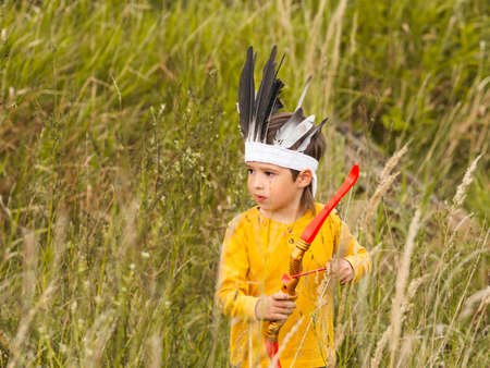 Little boy is playing American Indian on field. Kid has handmade headdress made of feathers and bow with arrows. Costume role play. Outdoor leisure activity. Fall season.の写真素材