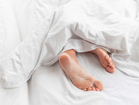 View on bare feet under blanket. Bedroom lit with morning light. Woman sleeps with comfort. White crumpled linen.の写真素材