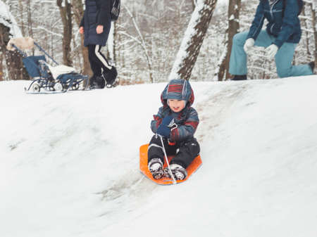 Kid is sledding down the hill. Mother helps boy to push off and ride down through snow. Winter fun. Family time outdoors. Active recreation. Leisure activity in cold season.の写真素材