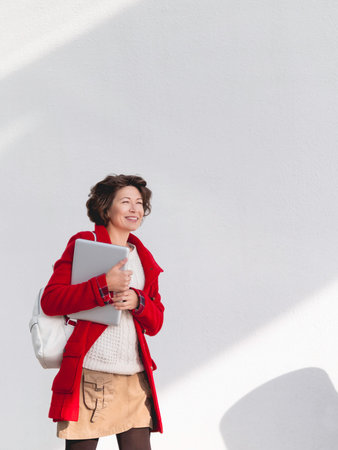 Curly woman in red coat is walking by white wall. Smiling student with laptop. Freelancer with portable device for work. Modern lifestyle.の写真素材