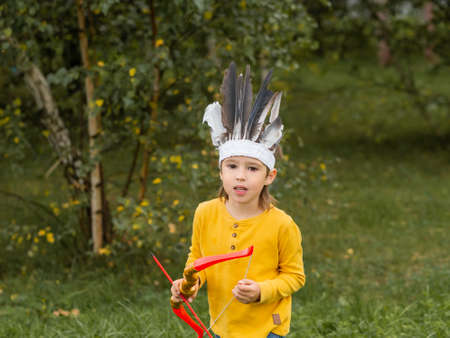 Little boy is playing American Indian on field. Kid has handmade headdress made of feathers and bow with arrows. Costume role play. Outdoor leisure activity. Fall season.の写真素材