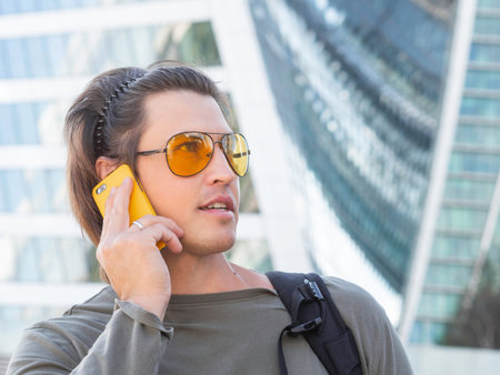 Young man with yellow eyeglasses speaks by smartphone on big city background. Modern buildings with...