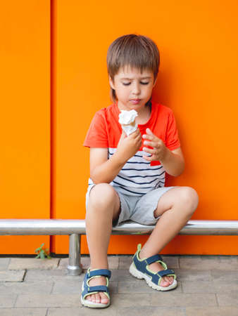 Little boy eats ice cream outdoors. Kid on monochrome, bright orange wall background. Tasty cold dessert at summer.の写真素材