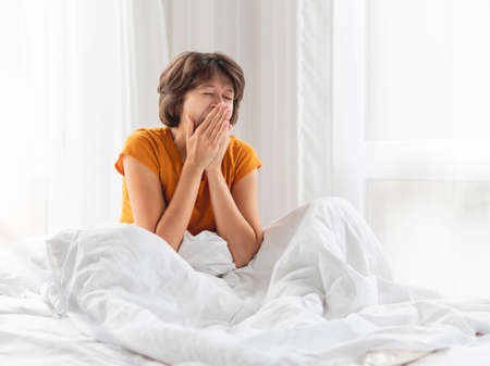 Sleepy woman is yawning in bed, covered with white blanket. It's hard to wake up early in morning.の写真素材