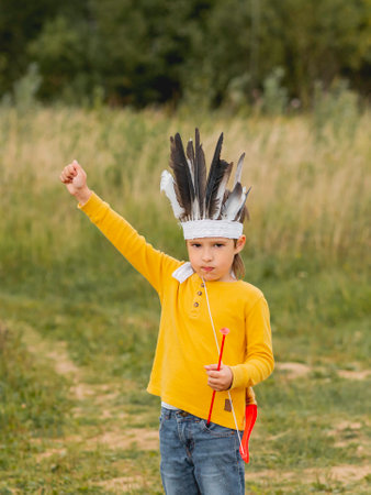 Little boy is playing American Indian on field. Kid has handmade headdress made of feathers and bow with arrows. Costume role play. Outdoor leisure activity. Fall season.の写真素材