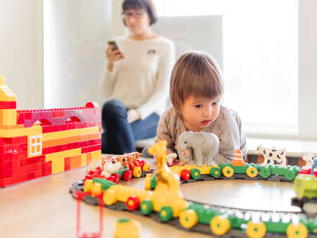Toddler plays with colorful toy blocks while his mother or babysitter texting in smartphone. Little boy stares on toy constructor. Interior of kindergarten or nursery.の写真素材